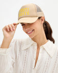 A woman wearing a beige trucker-style cap with yellow embroidered text reading 'TOUT DE SUITE' and a white lace blouse, photographed against a clean white background.