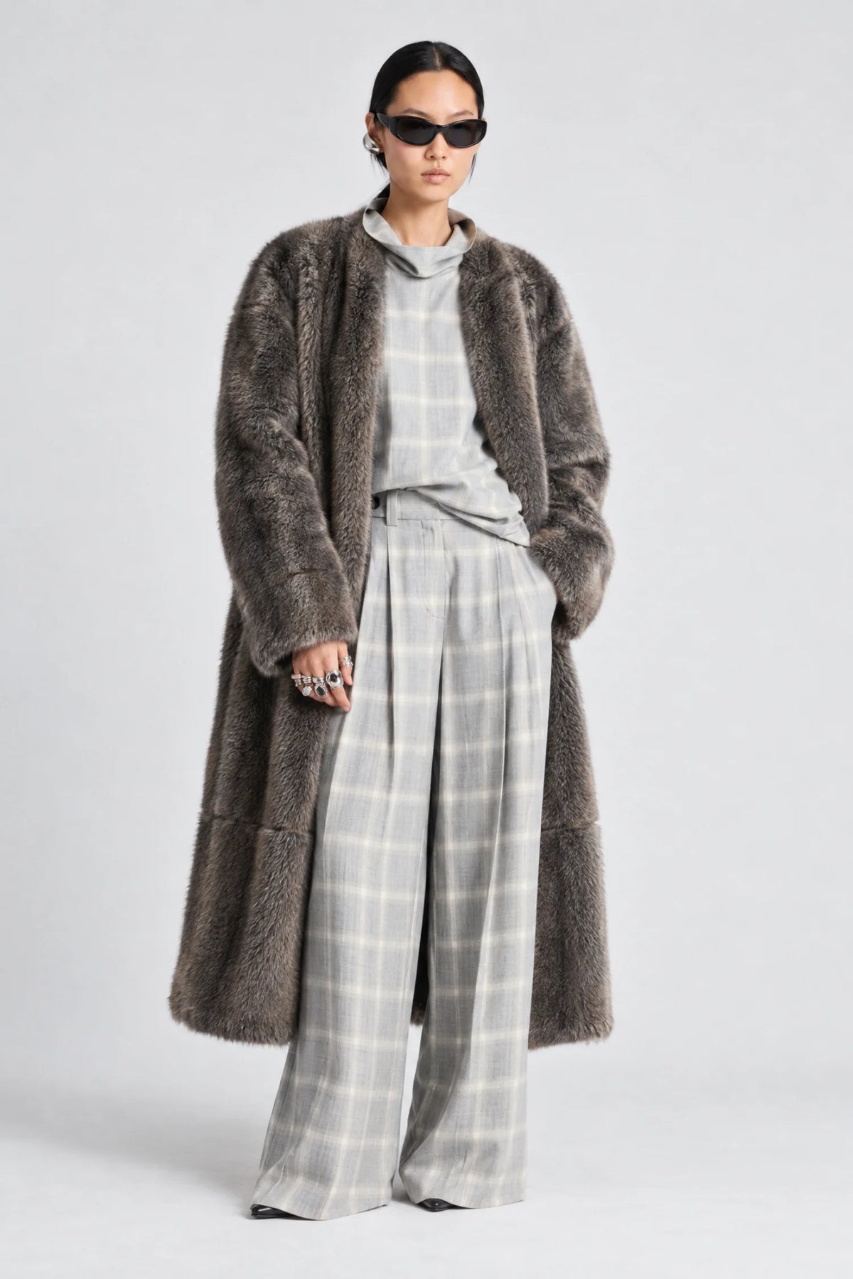 A model stands against a light gray studio background wearing a long, dark brown shaggy faux-fur coat over a light gray plaid shirt and matching wide-leg trousers.