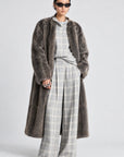 A model stands against a light gray studio background wearing a long, dark brown shaggy faux-fur coat over a light gray plaid shirt and matching wide-leg trousers.