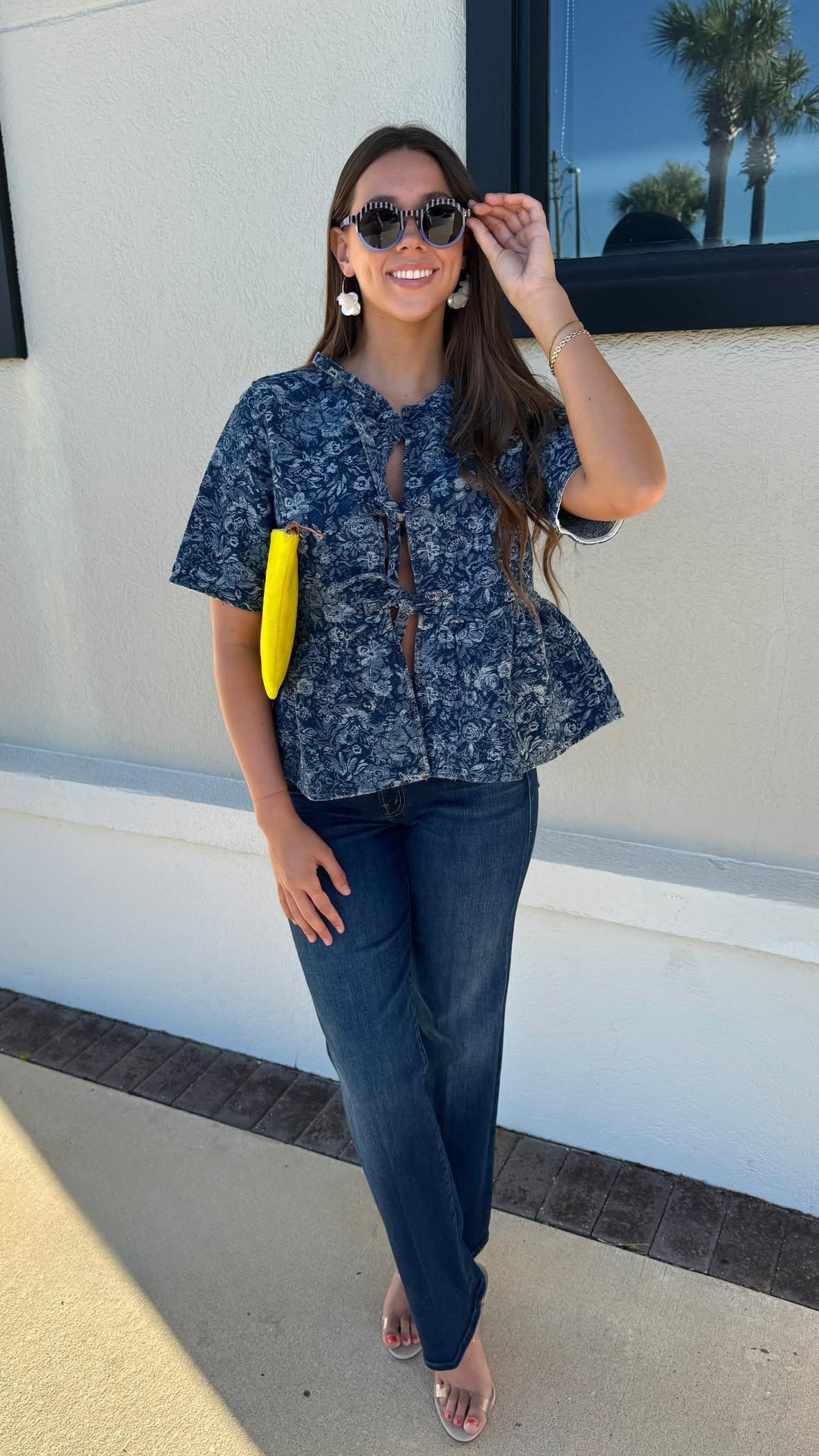 Woman in a blue patterned top and jeans standing in front of a white wall.