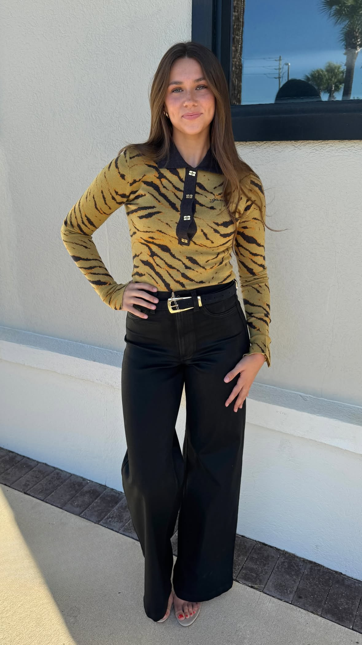 Woman wearing a tiger-striped sweater and black pants standing against a white wall.