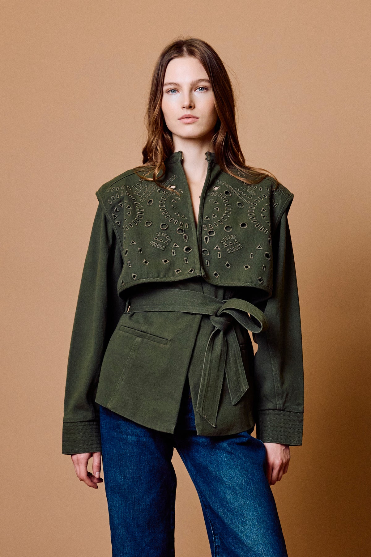A woman modeling an olive green cropped jacket with intricate metallic embroidery across the chest and a matching tie belt, paired with blue jeans against a solid tan studio background.