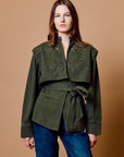 A woman modeling an olive green cropped jacket with intricate metallic embroidery across the chest and a matching tie belt, paired with blue jeans against a solid tan studio background.