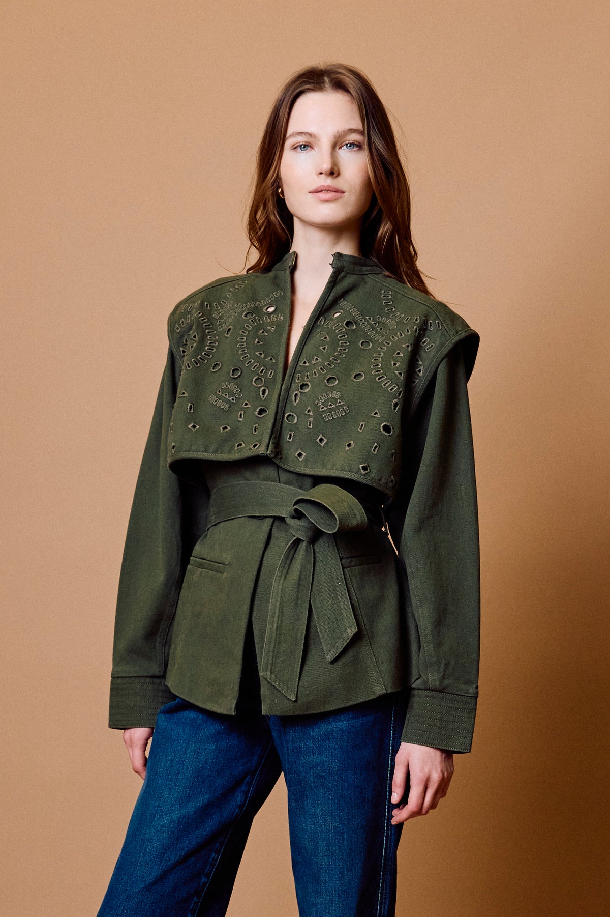 A woman modeling an olive green cropped jacket with intricate embroidery and a matching tie belt, paired with blue jeans, against a solid tan studio background.