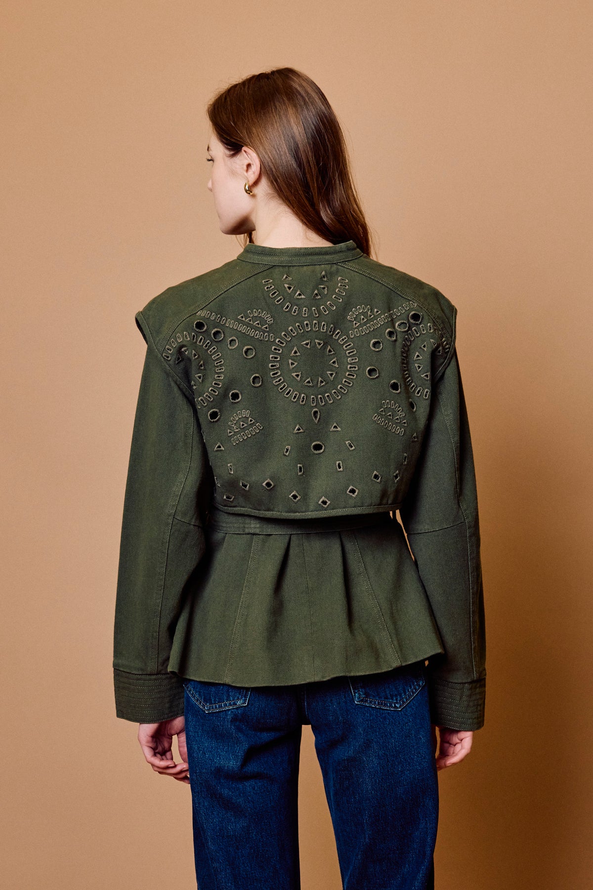 A woman viewed from the back wearing an olive green cropped jacket with decorative cutout embroidery on the back, paired with blue jeans, against a solid tan studio background.
