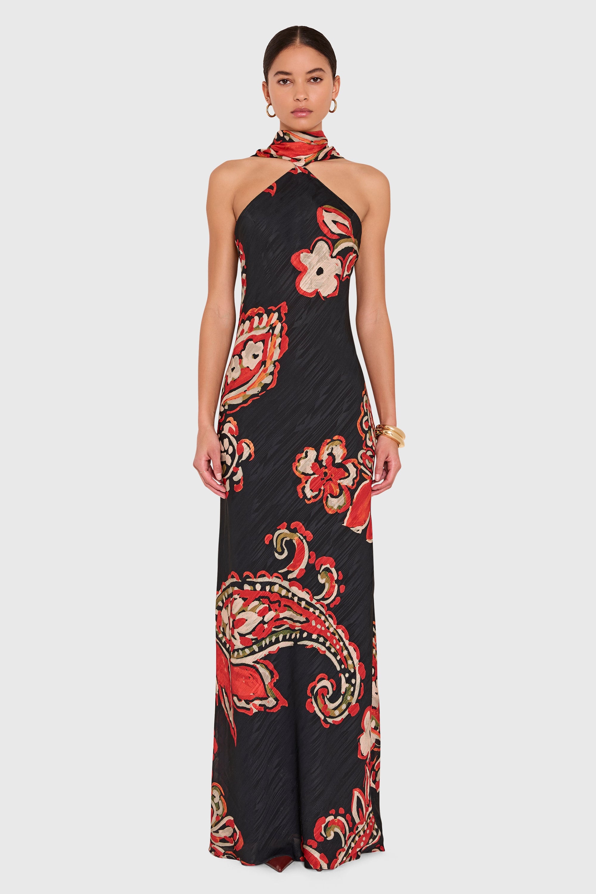 A model wearing a black halter-neck maxi dress with a red, orange, and white paisley and floral pattern, paired with a matching red neck scarf, against a light gray background.
