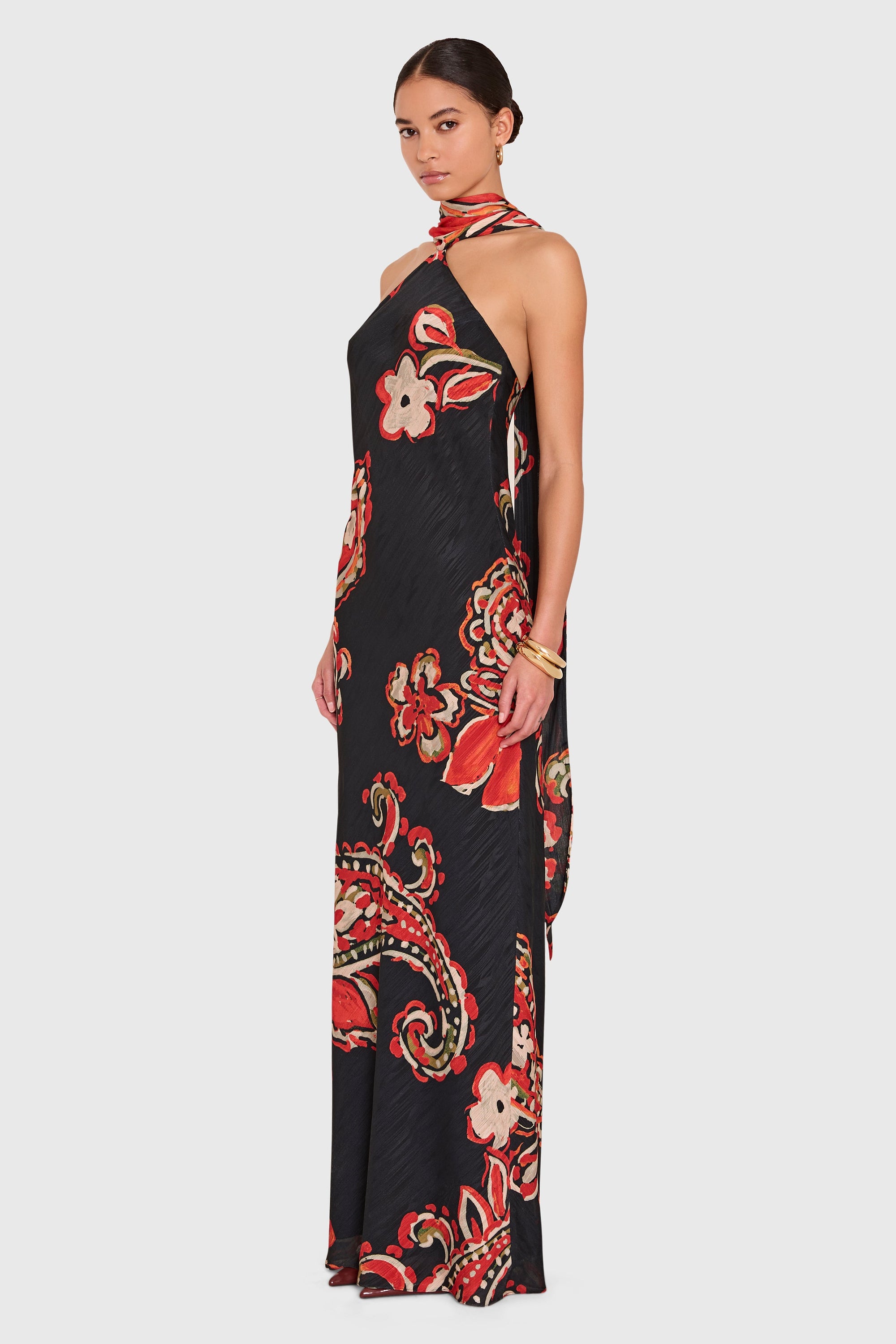 A woman modeling a black halter-neck maxi dress with a vibrant red, orange, and white paisley and floral print, set against a solid light gray background.