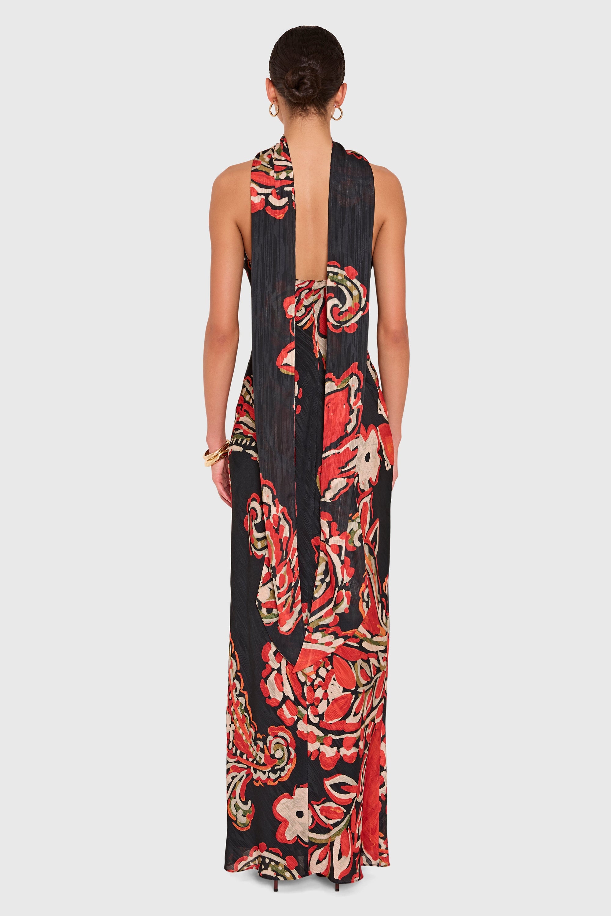 A woman viewed from the back wearing a sleeveless, floor-length halter dress with a bold floral print in red, orange, cream, and black on a light gray studio background.