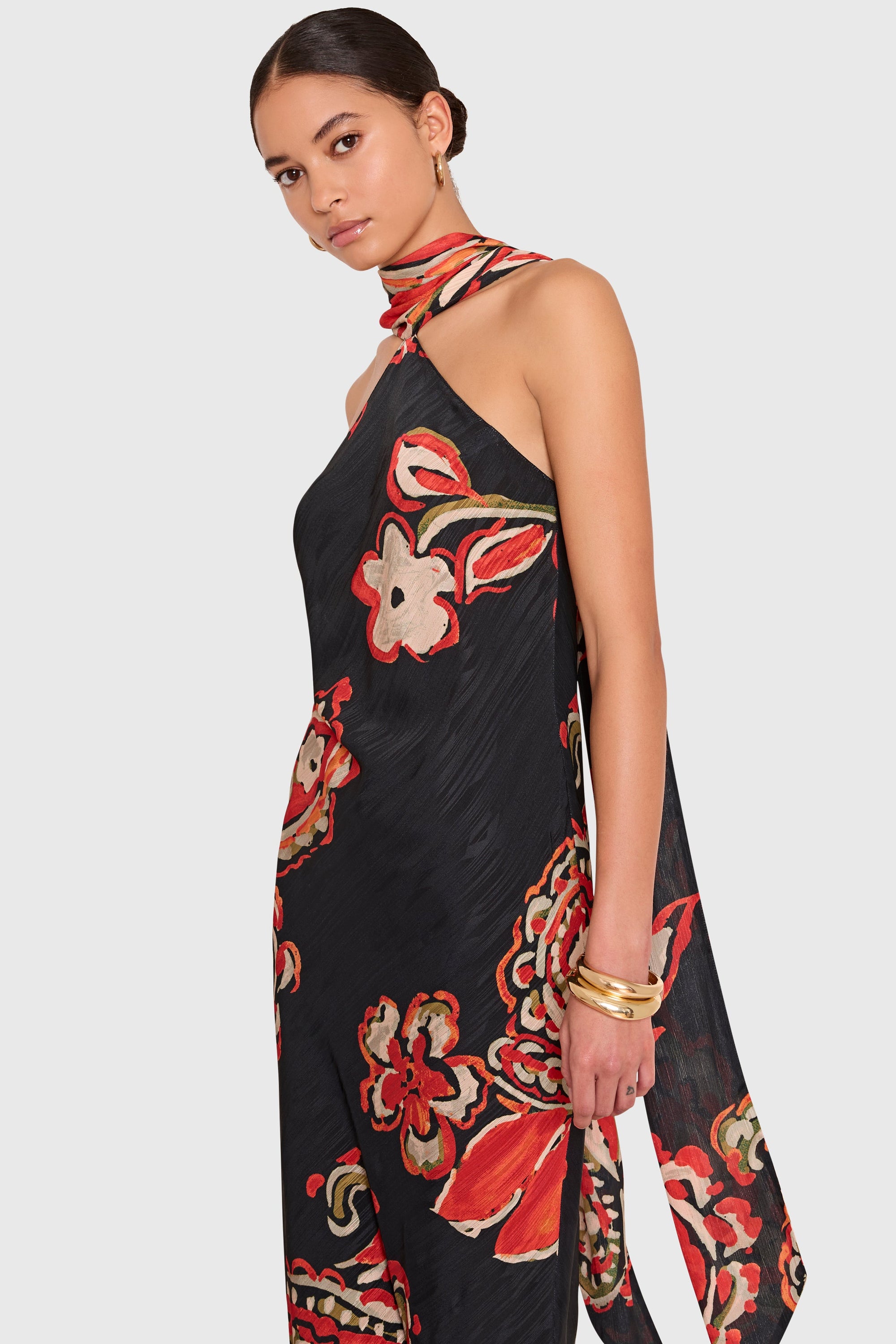 A woman modeling a black halter-neck maxi dress with a vibrant red, orange, and beige floral print, set against a solid light gray background.