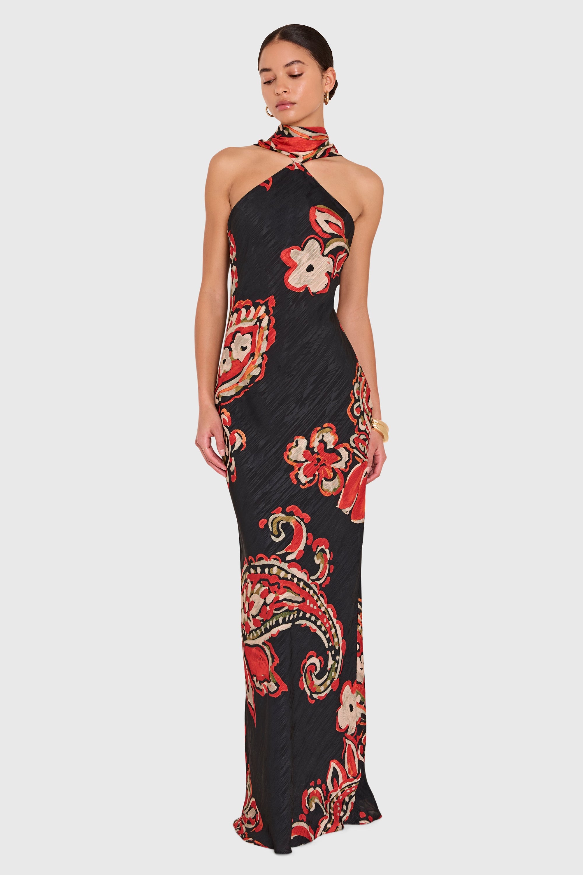 A woman modeling a sleeveless halter-neck maxi dress with a black base and a vibrant red, beige, and green paisley floral pattern, set against a solid light gray background.