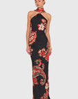 A woman modeling a sleeveless halter-neck maxi dress with a black base and a vibrant red, beige, and green paisley floral pattern, set against a solid light gray background.