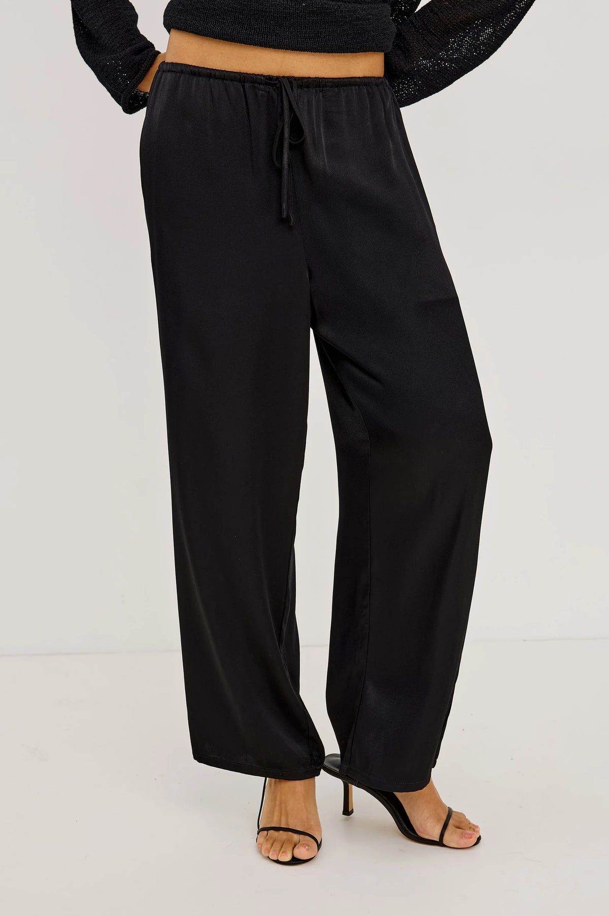 Black wide-leg drawstring pants shown on a model from the waist down, paired with black strappy high-heeled sandals against a light gray studio background.