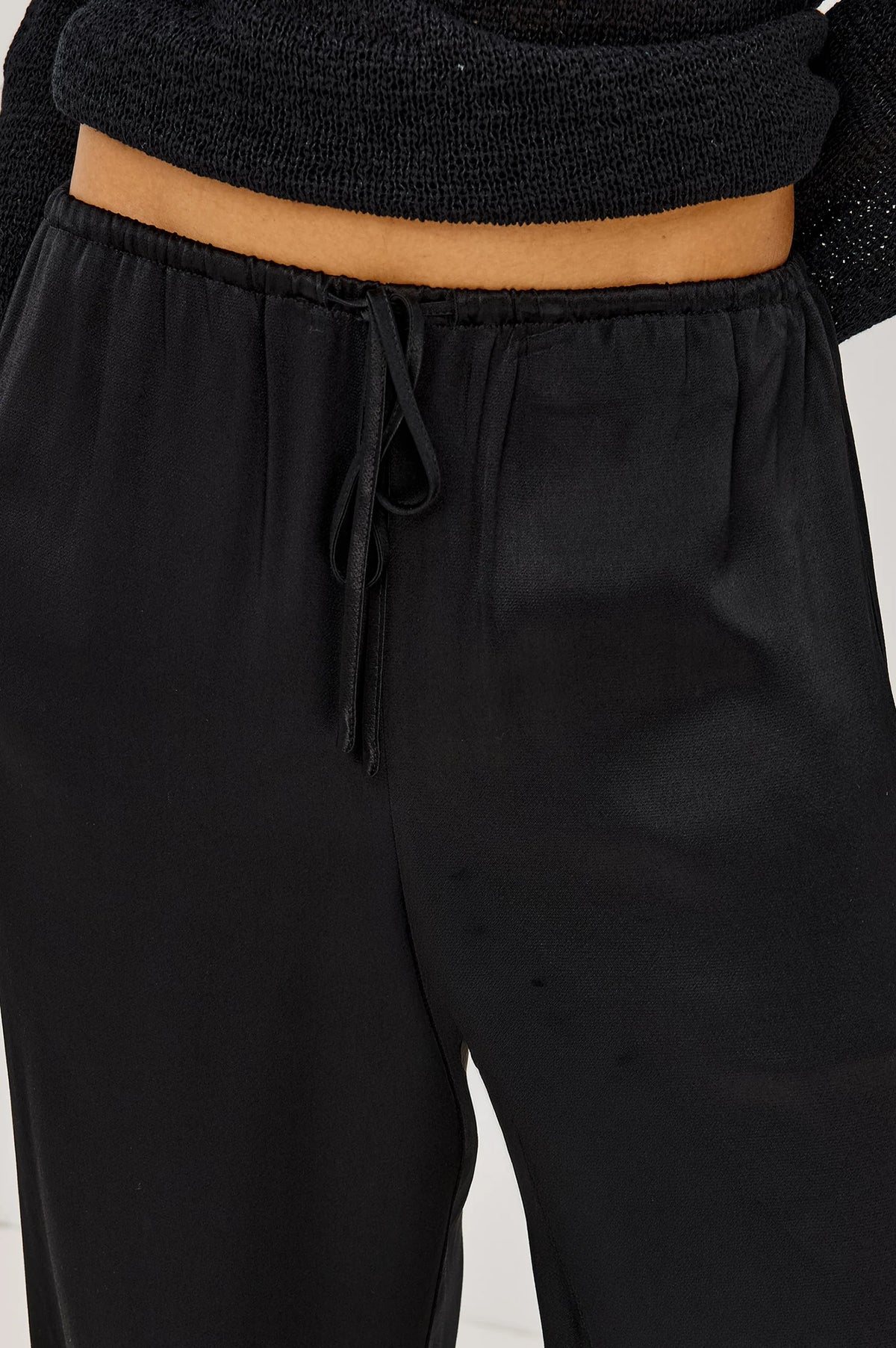 Close-up of a person wearing black drawstring sweatpants and a black ribbed cropped top, showing the waistband and fabric texture against a light neutral background.