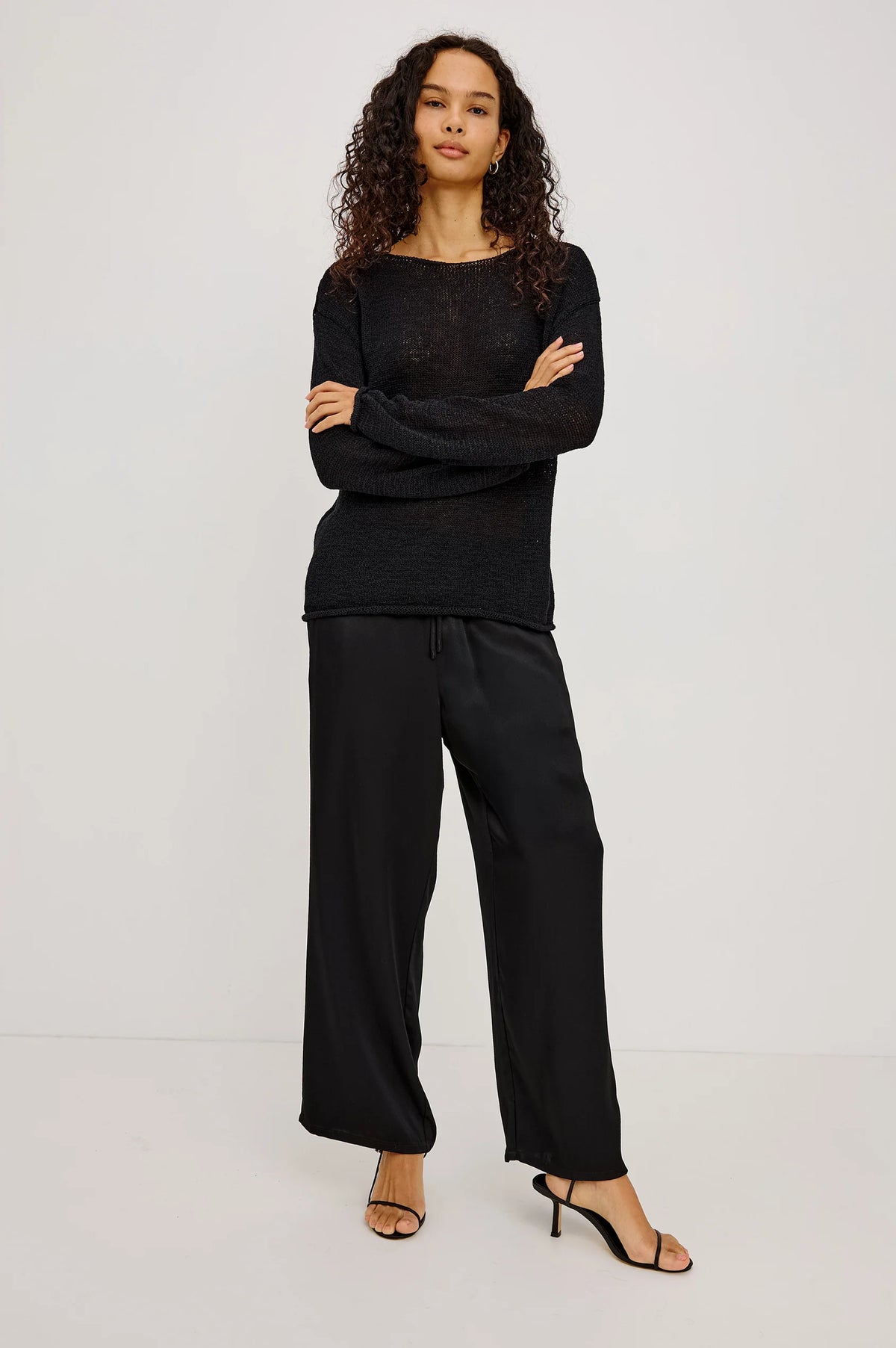 A model wearing a black long-sleeve knit top and matching black wide-leg trousers, paired with black heeled sandals, standing against a plain light gray studio background.