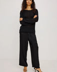 A model wearing a black long-sleeve knit top and matching black wide-leg trousers, paired with black heeled sandals, standing against a plain light gray studio background.