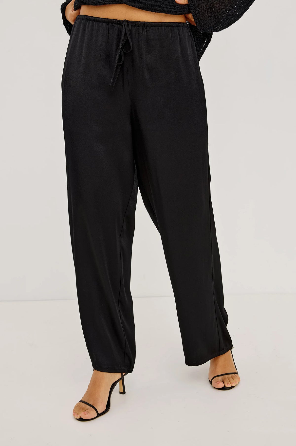 Black wide-leg drawstring pants shown on a model from the waist down, paired with black heeled sandals against a plain light gray studio background.