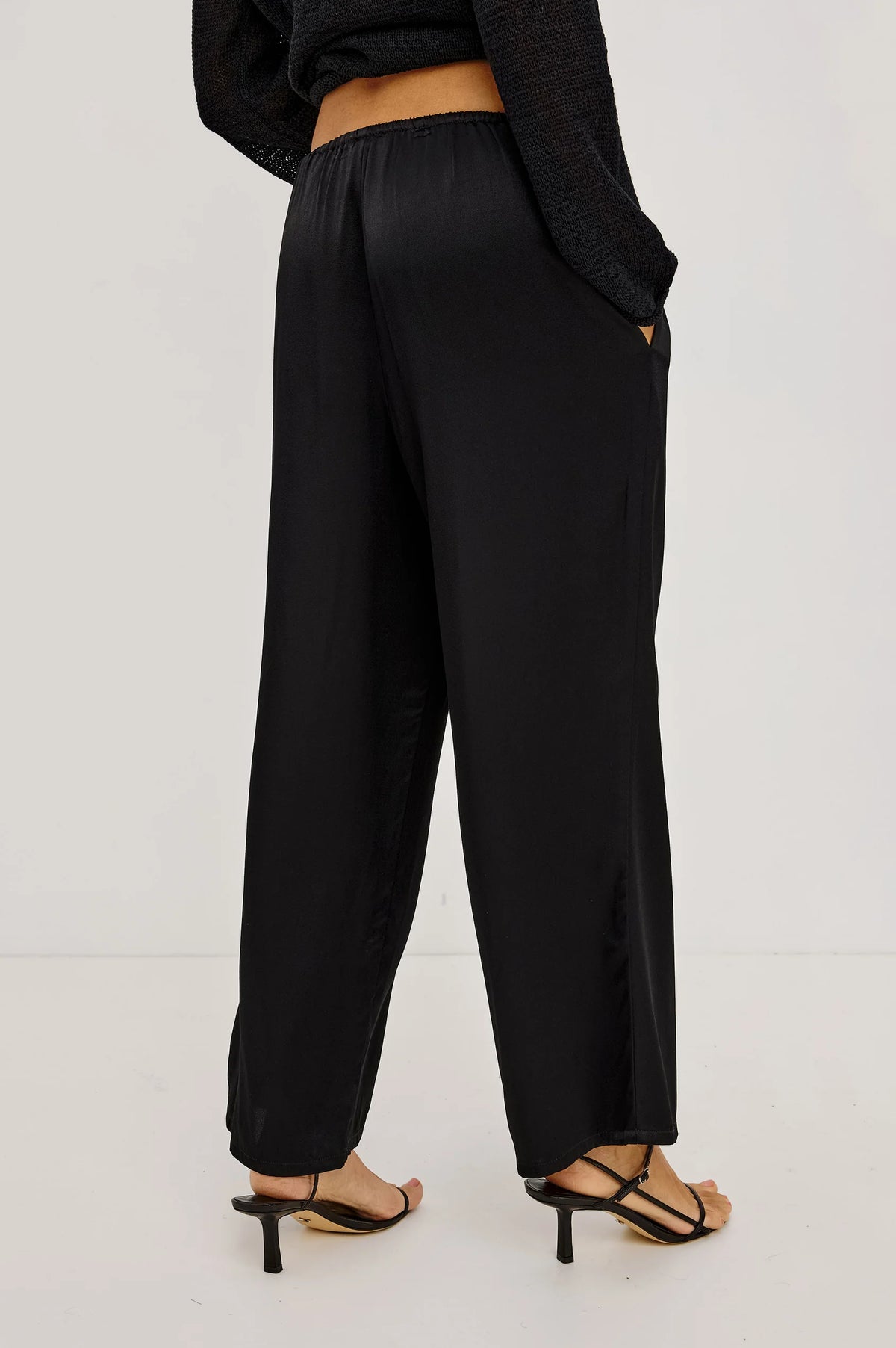 Black wide-leg trousers shown from the back on a model, paired with black strappy high-heeled sandals against a plain white studio background.