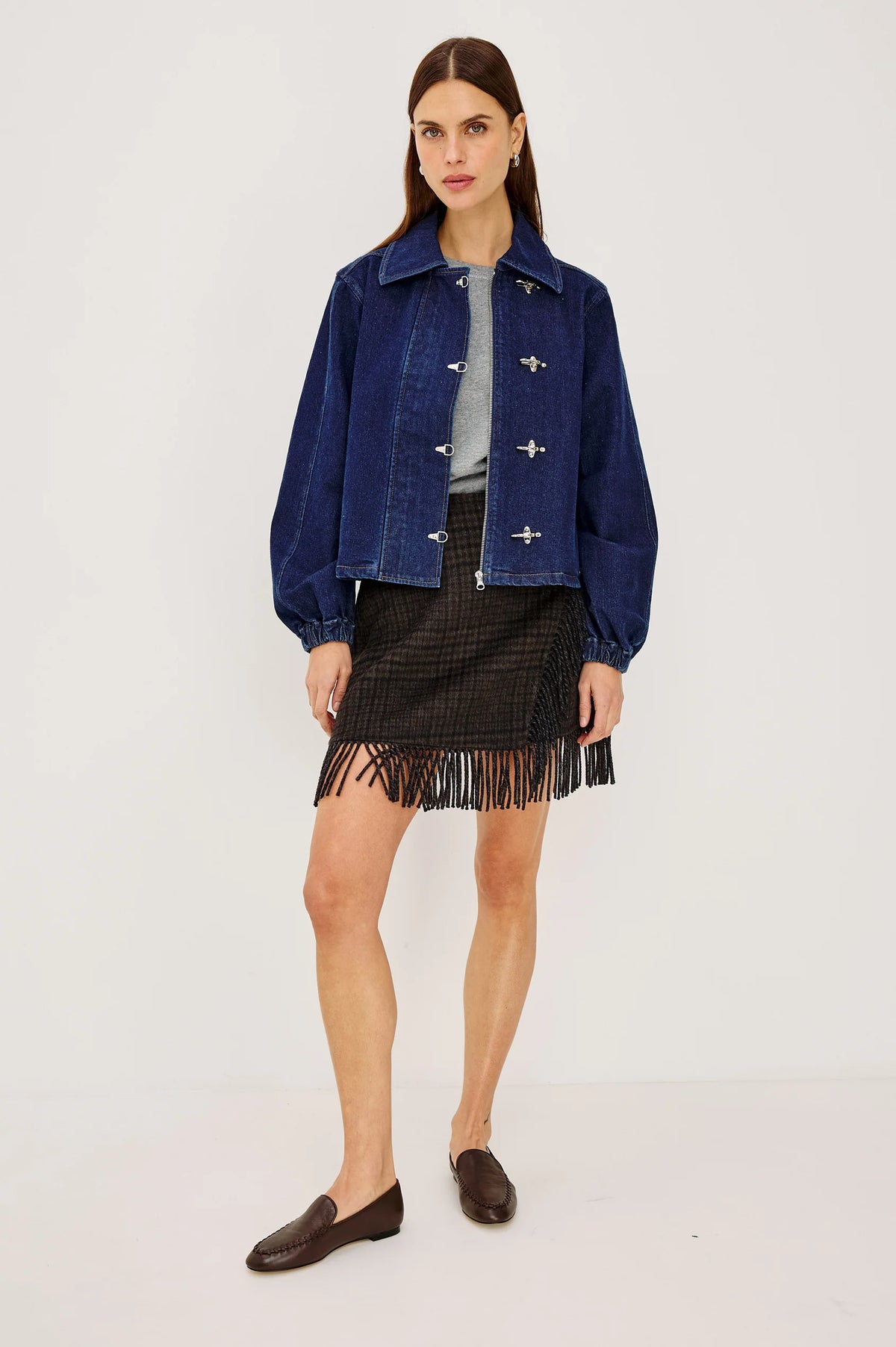 model wearing a fringe plaid skirt with a grey top and jacket, loafers, on a light grey background 