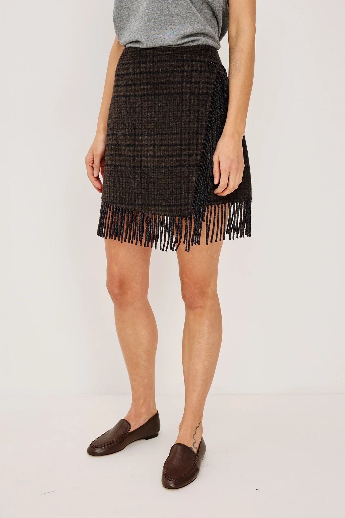 model wearing a fringe plaid skirt with a grey top, loafers, on a light grey background 