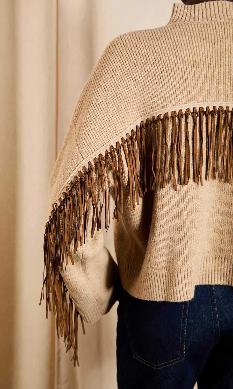 A close-up of a beige knit sweater with a wide brown fringe trim across the shoulders, worn over dark blue jeans against a soft beige curtain background.