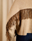 A close-up of a beige knit sweater with a wide brown fringe trim across the shoulders, worn over dark blue jeans against a soft beige curtain background.