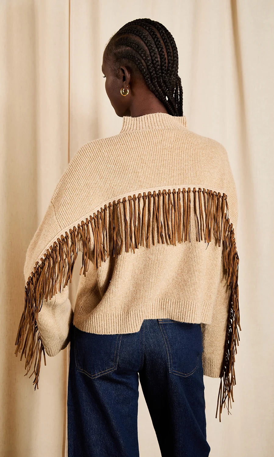 A model shown from the back wearing a cream-colored knit poncho sweater with a wide brown faux-suede fringe detail across the shoulders, paired with dark blue jeans, set against a neutral beige curtain backdrop.