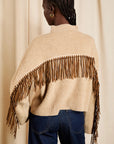 A model shown from the back wearing a cream-colored knit poncho sweater with a wide brown faux-suede fringe detail across the shoulders, paired with dark blue jeans, set against a neutral beige curtain backdrop.