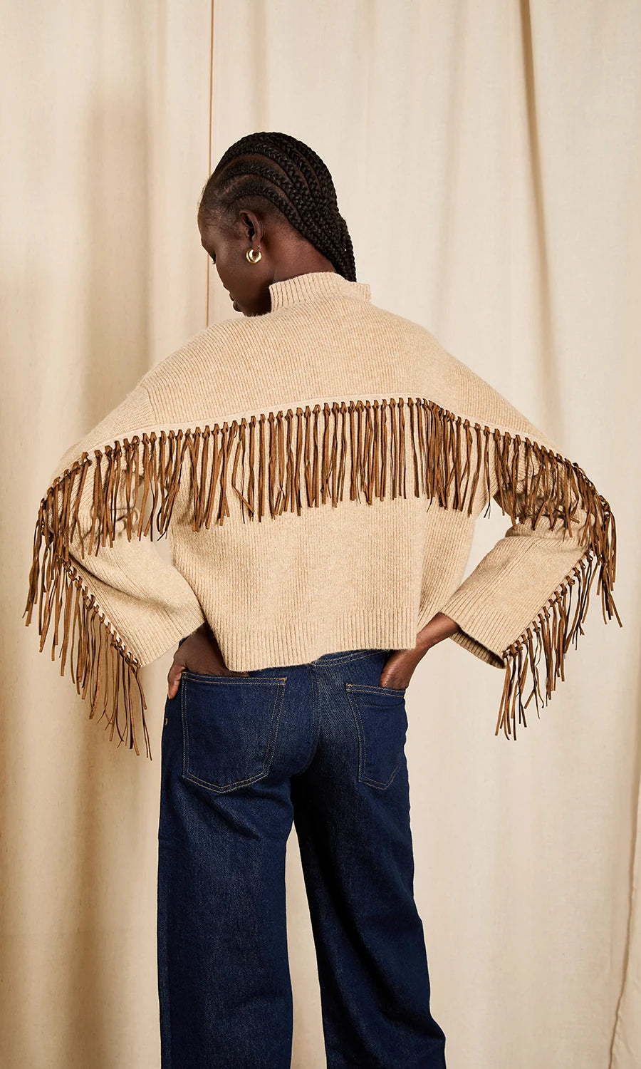 A model shown from behind wearing a cream-colored knit poncho sweater with brown fringe across the shoulders and blue jeans, standing against a neutral beige curtain backdrop.