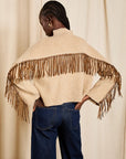 A model shown from behind wearing a cream-colored knit poncho sweater with brown fringe across the shoulders and blue jeans, standing against a neutral beige curtain backdrop.