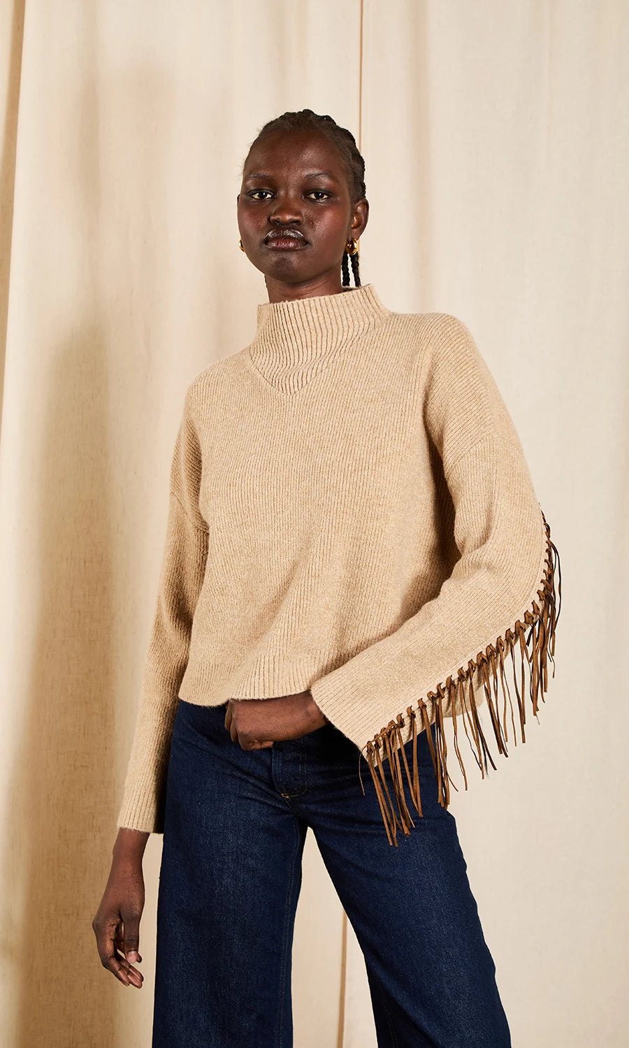 A model wearing a beige knit turtleneck sweater with long fringe detailing on the sleeves, paired with dark blue jeans, standing against a neutral beige curtain backdrop.