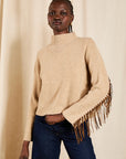 A model wearing a beige knit turtleneck sweater with long fringe detailing on the sleeves, paired with dark blue jeans, standing against a neutral beige curtain backdrop.