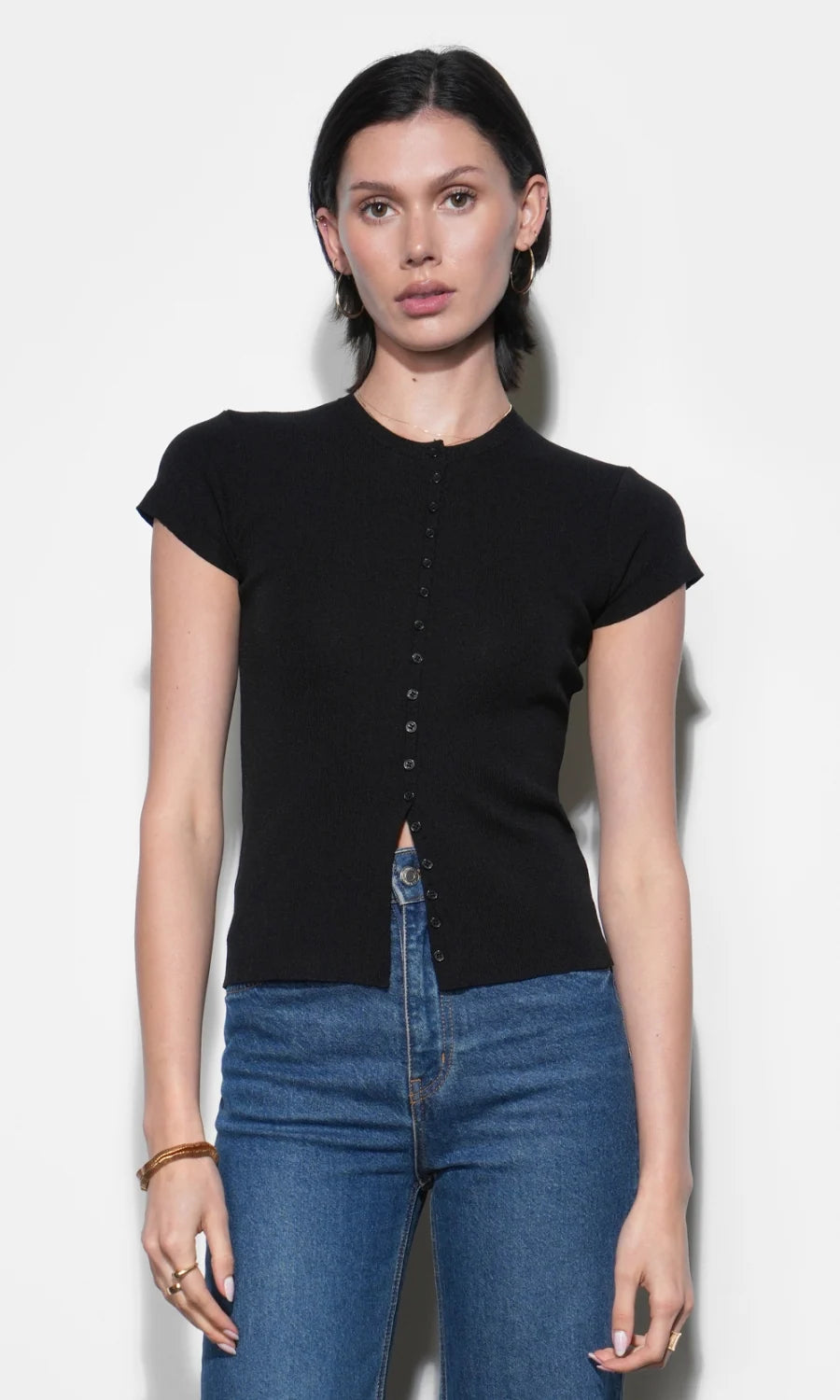 A woman modeling a black short-sleeve knit top with a button-down front and a small front cutout, paired with blue jeans, against a plain light background.
