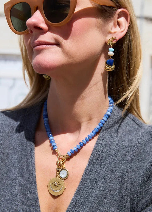 A woman wearing a blue beaded necklace with gold-tone charms and matching drop earrings, photographed outdoors against a blurred light-colored building background.