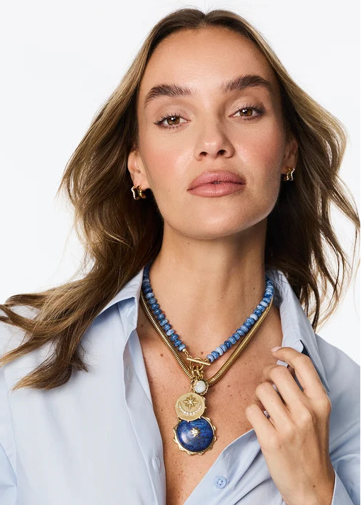 A woman with light brown hair models a gold-tone layered necklace featuring a blue beaded collar and a large blue and gold pendant, paired with a light blue button-down shirt against a white studio background.