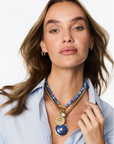 A woman with light brown hair models a gold-tone layered necklace featuring a blue beaded collar and a large blue and gold pendant, paired with a light blue button-down shirt against a white studio background.