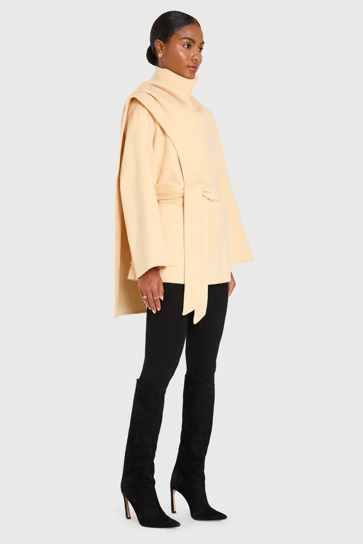 Woman wearing a beige coat, black pants, and high-heeled shoes on a white background side view