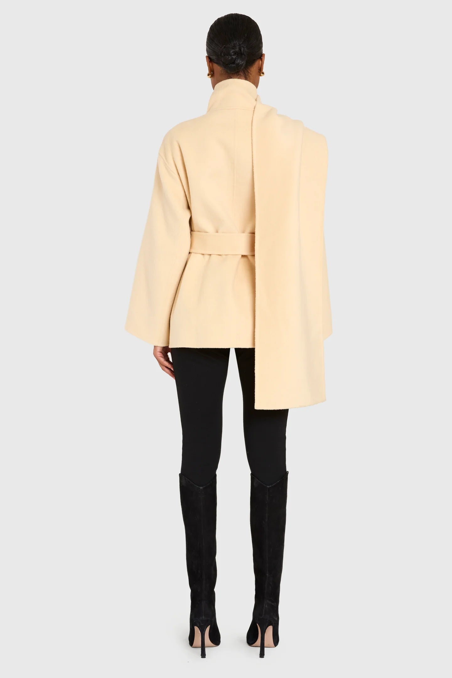 Person wearing a light yellow coat with a belt, black pants, and high heels on a white background back view