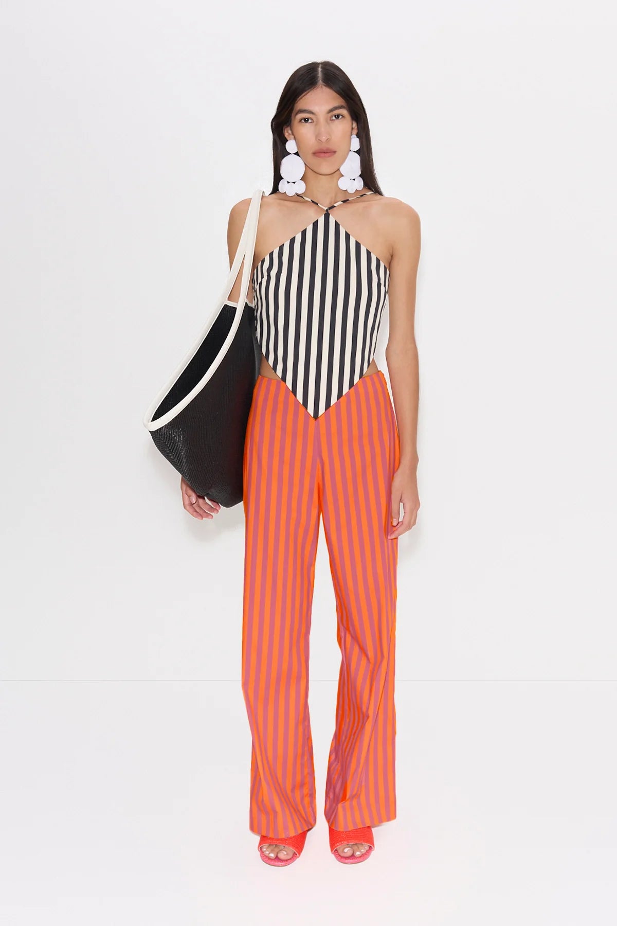 Woman wearing a black and white striped top and orange pants, holding a black and white bag on a white background.