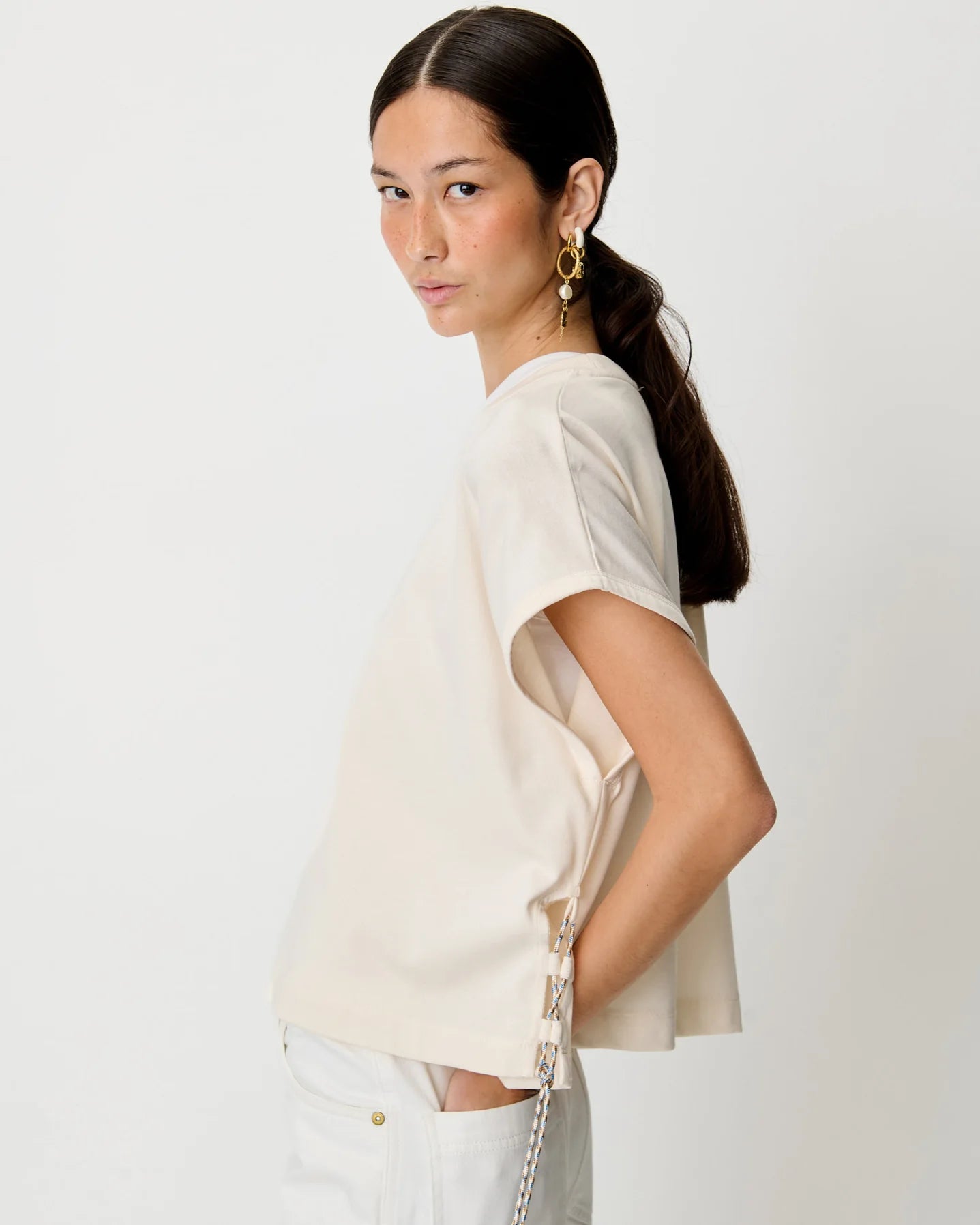 A woman with her hair in a ponytail models a short-sleeve cream-colored top with side lace-up detailing, paired with white pants, against a plain light gray background.