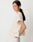 A woman with her hair in a ponytail models a short-sleeve cream-colored top with side lace-up detailing, paired with white pants, against a plain light gray background.
