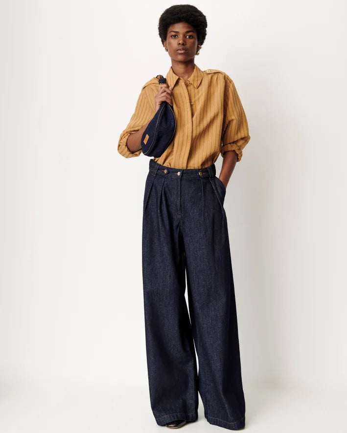 A model stands against a plain light background wearing a mustard yellow striped button-up shirt, dark wide-leg denim pants, and carrying a dark blue shoulder bag.