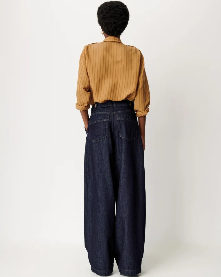 A person shown from the back wearing a mustard yellow vertically striped long-sleeve shirt and dark wide-leg denim jeans against a plain light gray studio background.