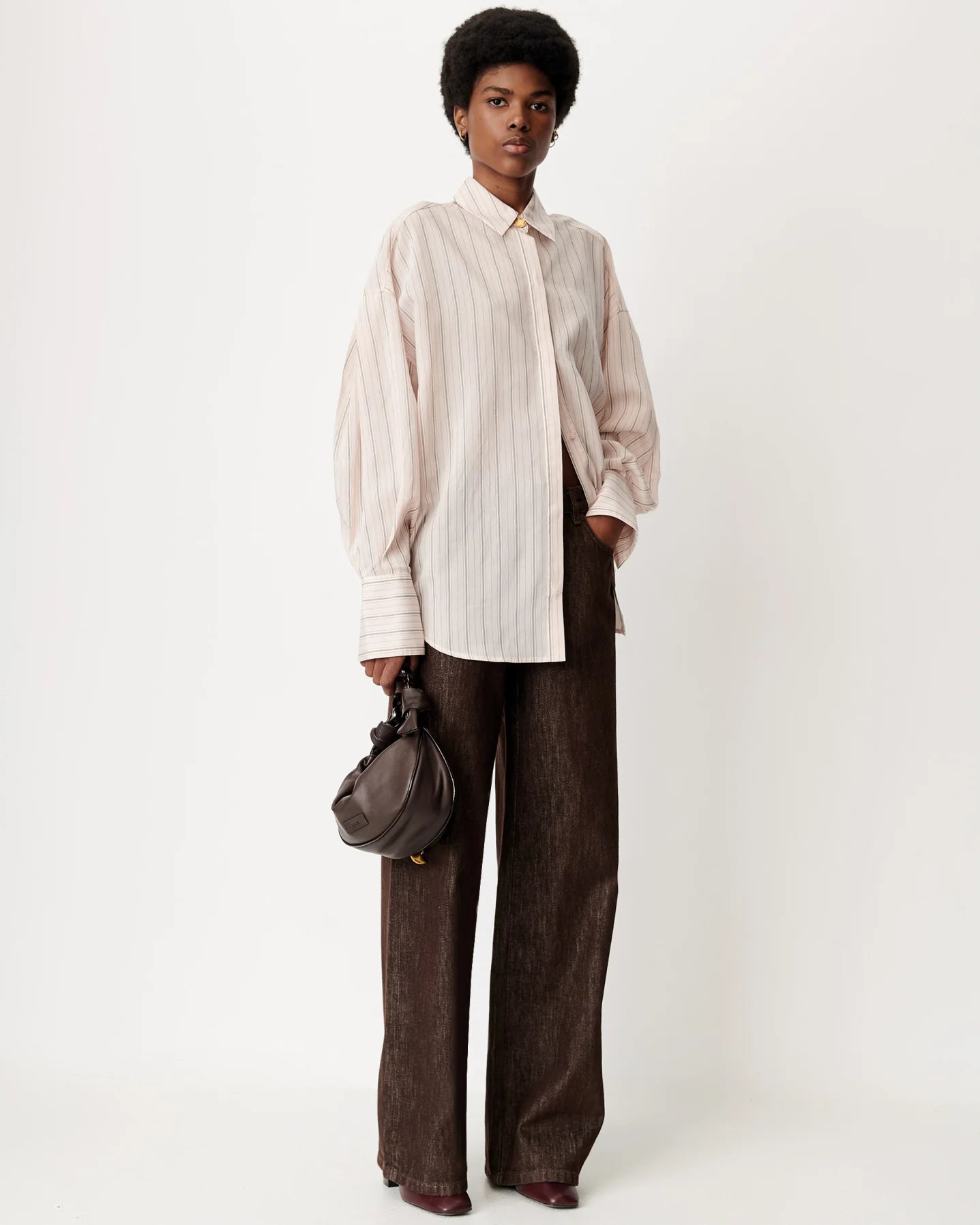 A model stands against a plain light background wearing a cream and beige vertically striped button-up shirt with voluminous sleeves, dark brown wide-leg trousers, and holding a small dark brown shoulder bag.