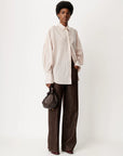 A model stands against a plain light background wearing a cream and beige vertically striped button-up shirt with voluminous sleeves, dark brown wide-leg trousers, and holding a small dark brown shoulder bag.