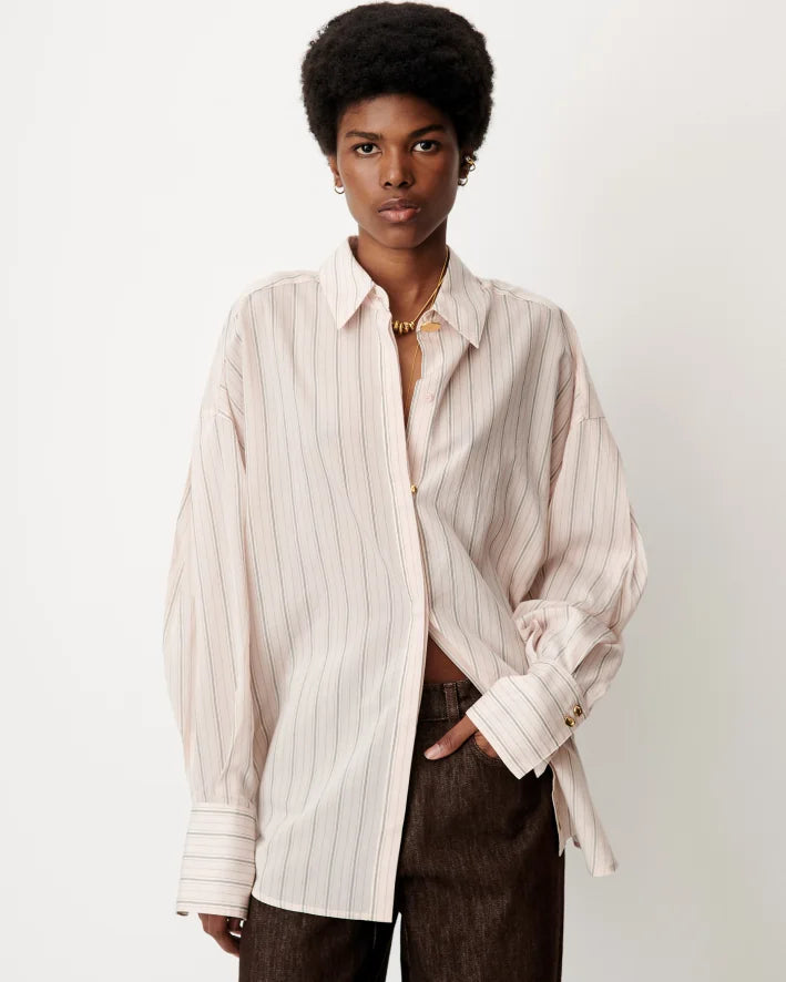 A model wearing an oversized, long-sleeve button-up shirt with thin vertical stripes in cream and light brown, paired with dark brown trousers, against a plain off-white background.