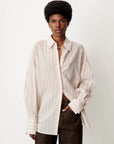 A model wearing an oversized, long-sleeve button-up shirt with thin vertical stripes in cream and light brown, paired with dark brown trousers, against a plain off-white background.