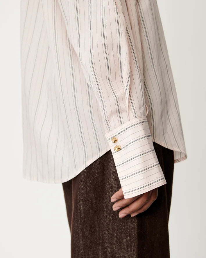 Close-up of a person wearing a cream and gray vertically striped shirt with gold buttons on the cuff, paired with dark brown textured trousers, against a plain light background.