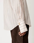 Close-up of a person wearing a cream and gray vertically striped shirt with gold buttons on the cuff, paired with dark brown textured trousers, against a plain light background.
