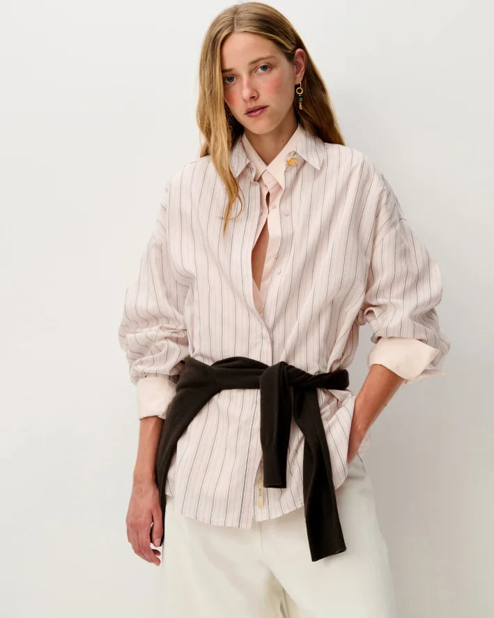 A woman modeling a light pink and white vertically striped button-down shirt with a black sweater around her waist, paired with white trousers, against a plain white studio background.