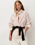 A woman modeling a light pink and white vertically striped button-down shirt with a black sweater around her waist, paired with white trousers, against a plain white studio background.